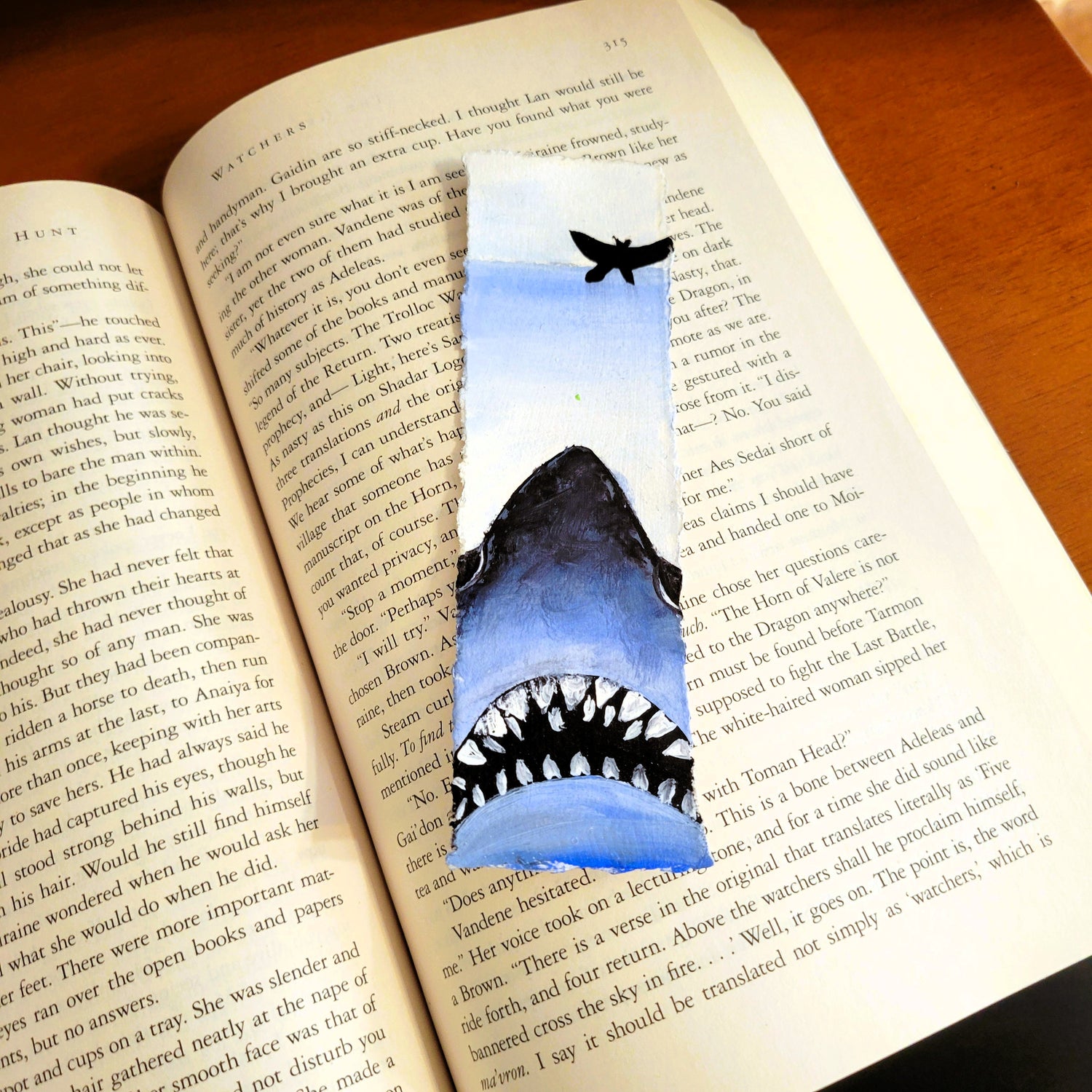 Hand-painted bookmark featuring a large blue-gray shark rising beneath a small black boat on dark water by Amy Bridges.
