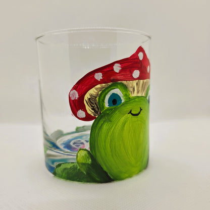 Hand-painted 11 oz highball glass featuring a whimsical frog sitting on a lily pad wearing a red mushroom hat with lily pad details wrapping around the glass.