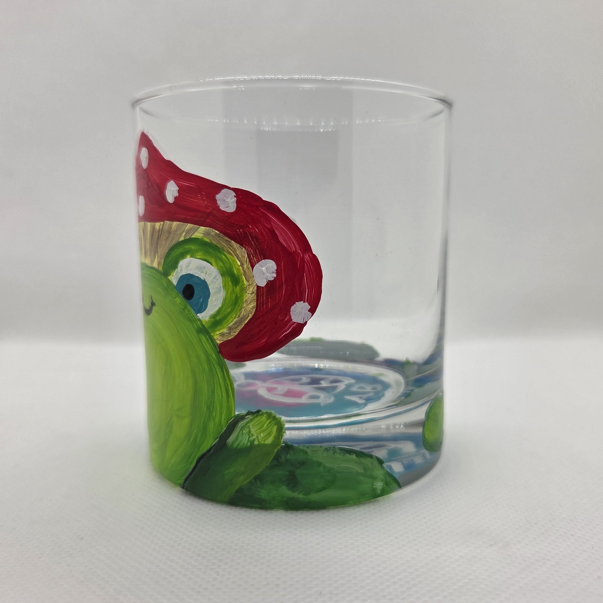 Hand-painted 11 oz highball glass featuring a whimsical frog sitting on a lily pad wearing a red mushroom hat with lily pad details wrapping around the glass.