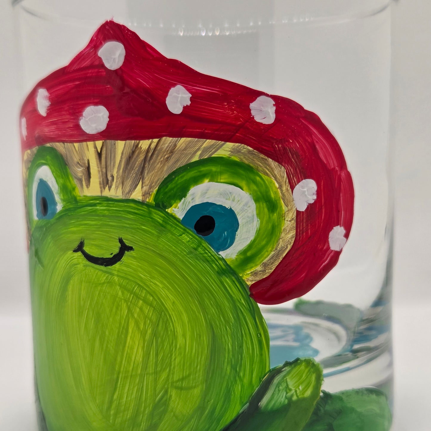 Hand-painted 11 oz highball glass featuring a whimsical frog sitting on a lily pad wearing a red mushroom hat with lily pad details wrapping around the glass.