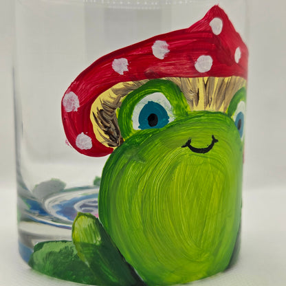 Hand-painted 11 oz highball glass featuring a whimsical frog sitting on a lily pad wearing a red mushroom hat with lily pad details wrapping around the glass.